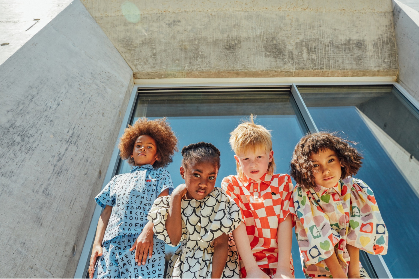 Four children standing in front of a modern building with large glass windows.